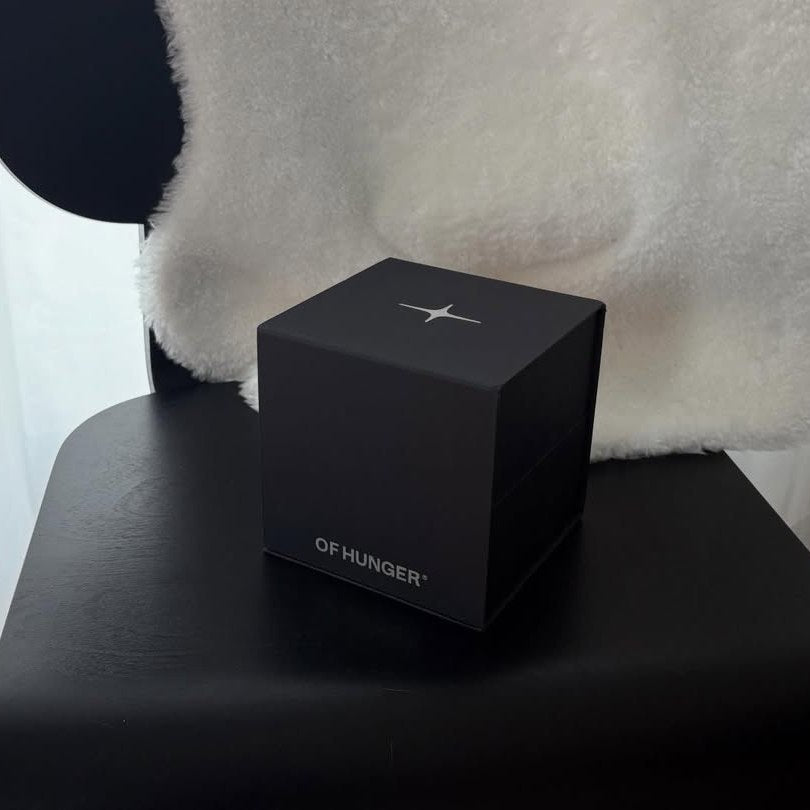 A black OF HUNGER presentation box on a chair beside a light-colored cushion.