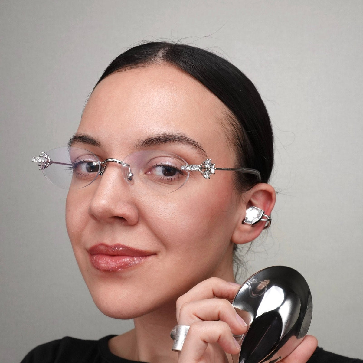 Electronic Gems worn with glasses while holding the closed silver-toned charging case.