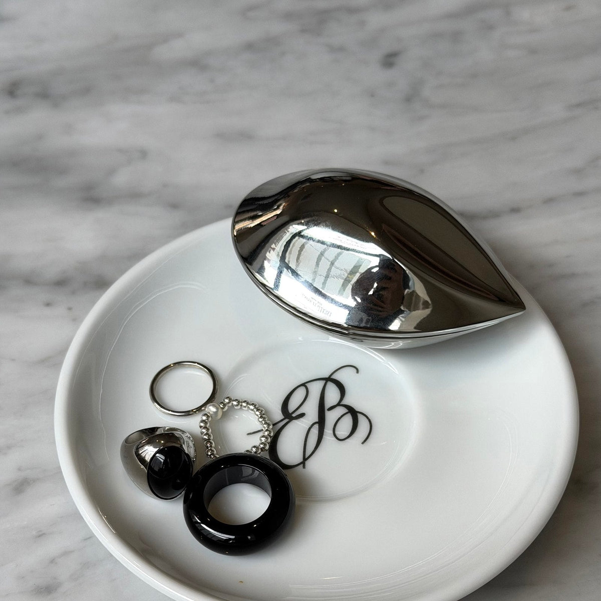 A closed silver-toned Electronic Gems charging case on a plate beside rings.